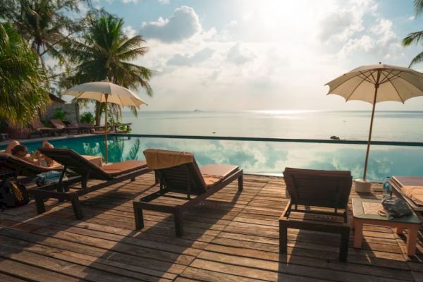A serene poolside scene with lounge chairs, umbrellas, and palm trees overlooking a calm sea during sunset.