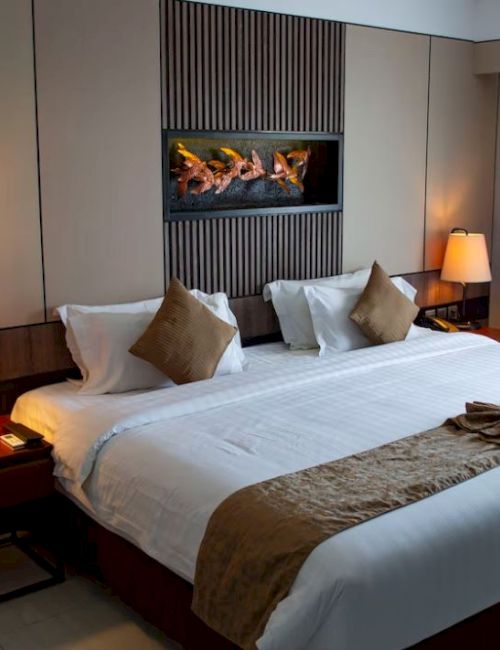 A neatly arranged hotel room features a king-sized bed, warm lighting, neutral decor, and a small fireplace on the wall.