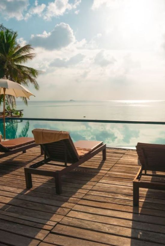 A serene beach scene with lounge chairs, umbrellas, and palm trees by a pool overlooking the ocean under a partly cloudy sky.