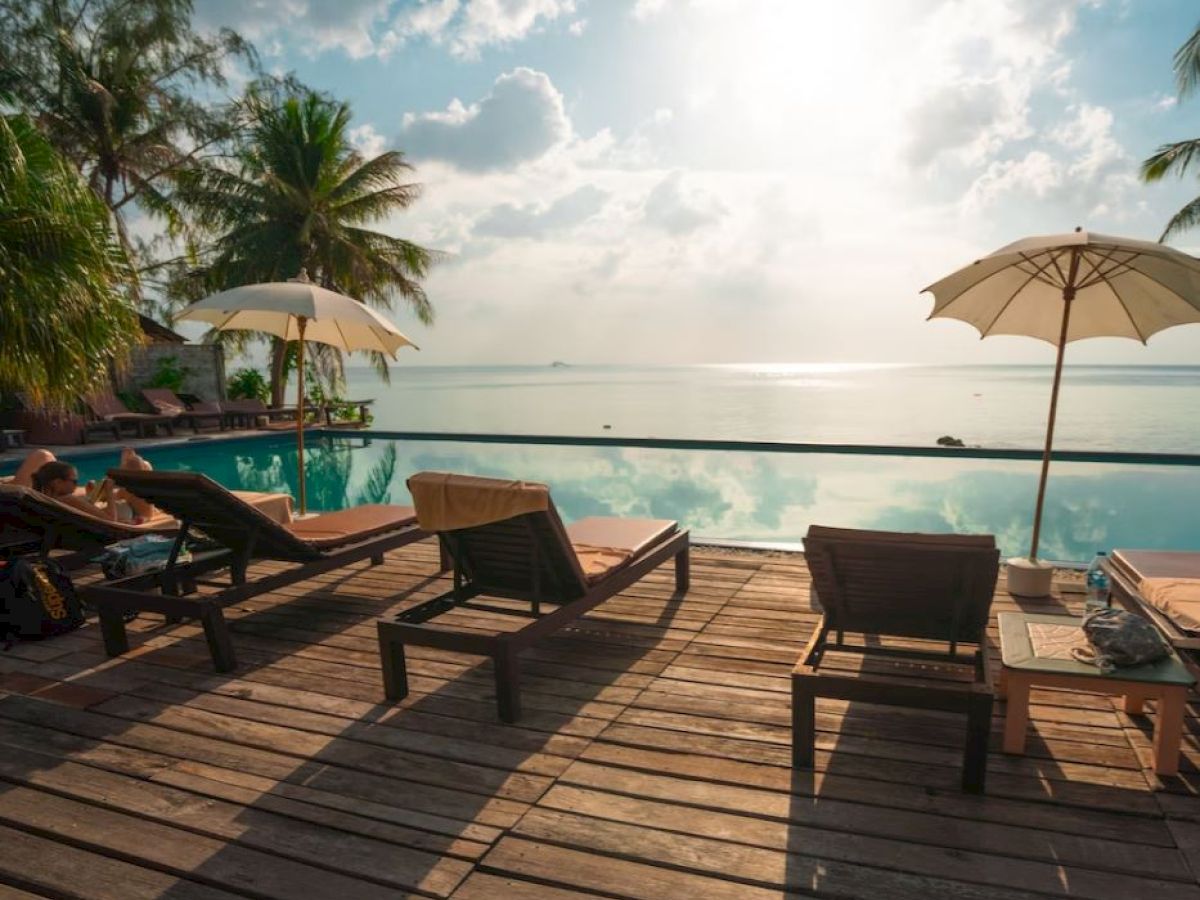 A serene poolside setting with loungers, umbrellas, palm trees, and a view of the ocean under a partly cloudy sky.