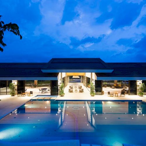 The image shows a luxurious, well-lit building with an expansive pool in the foreground under a vibrant blue evening sky.