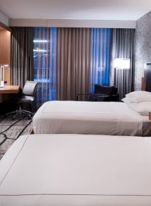The image shows a modern hotel room with two beds, a TV, desk, and decorative lighting, featuring a stylish wallpapered accent wall.