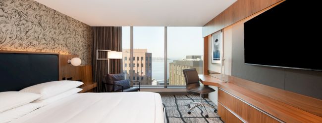 A modern hotel room with a bed, large TV, desk, and a window view of buildings and water.