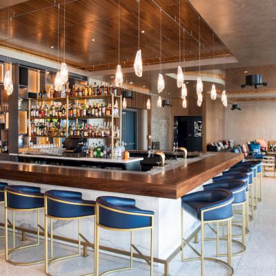 A modern bar with a central counter, blue stools, hanging lights, and a well-stocked shelf of bottles. Lounge seating is visible in the background.
