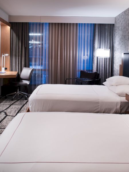 The image shows a modern hotel room with two beds, a desk, a TV, and stylish decor, featuring a patterned wall and floor.