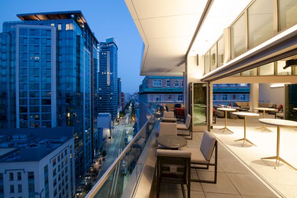A modern rooftop terrace with seating overlooks a city street lined with tall glass buildings during evening.