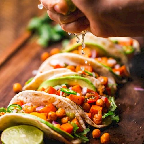 A hand squeezes lime over tacos filled with chickpeas, avocado, and vegetables, garnished with cilantro on a wooden board.