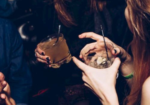 People are enjoying drinks in a lively setting, smiling and talking. Two hands hold cocktails, and there's a sense of fun and camaraderie.