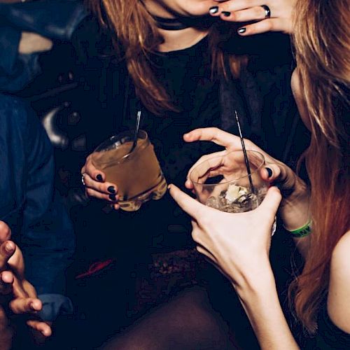People are enjoying drinks in a lively setting, smiling and talking. Two hands hold cocktails, and there's a sense of fun and camaraderie.