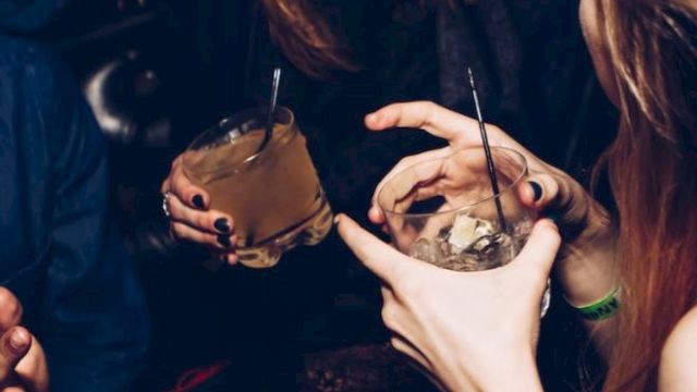 People are enjoying drinks in a lively setting, smiling and talking. Two hands hold cocktails, and there's a sense of fun and camaraderie.