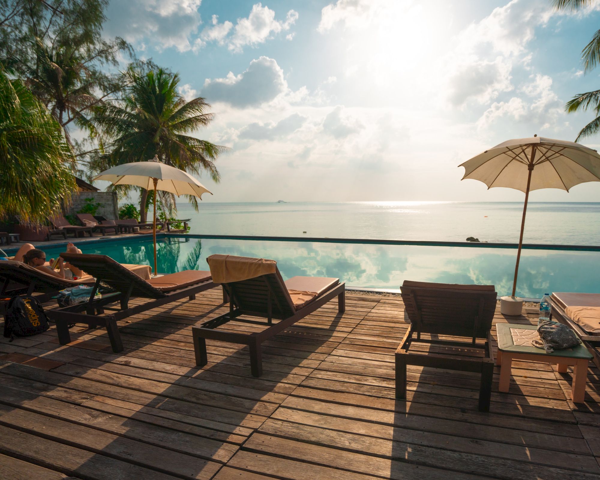 Poolside scene with deck chairs, umbrellas, and view of the ocean under a bright, cloudy sky.