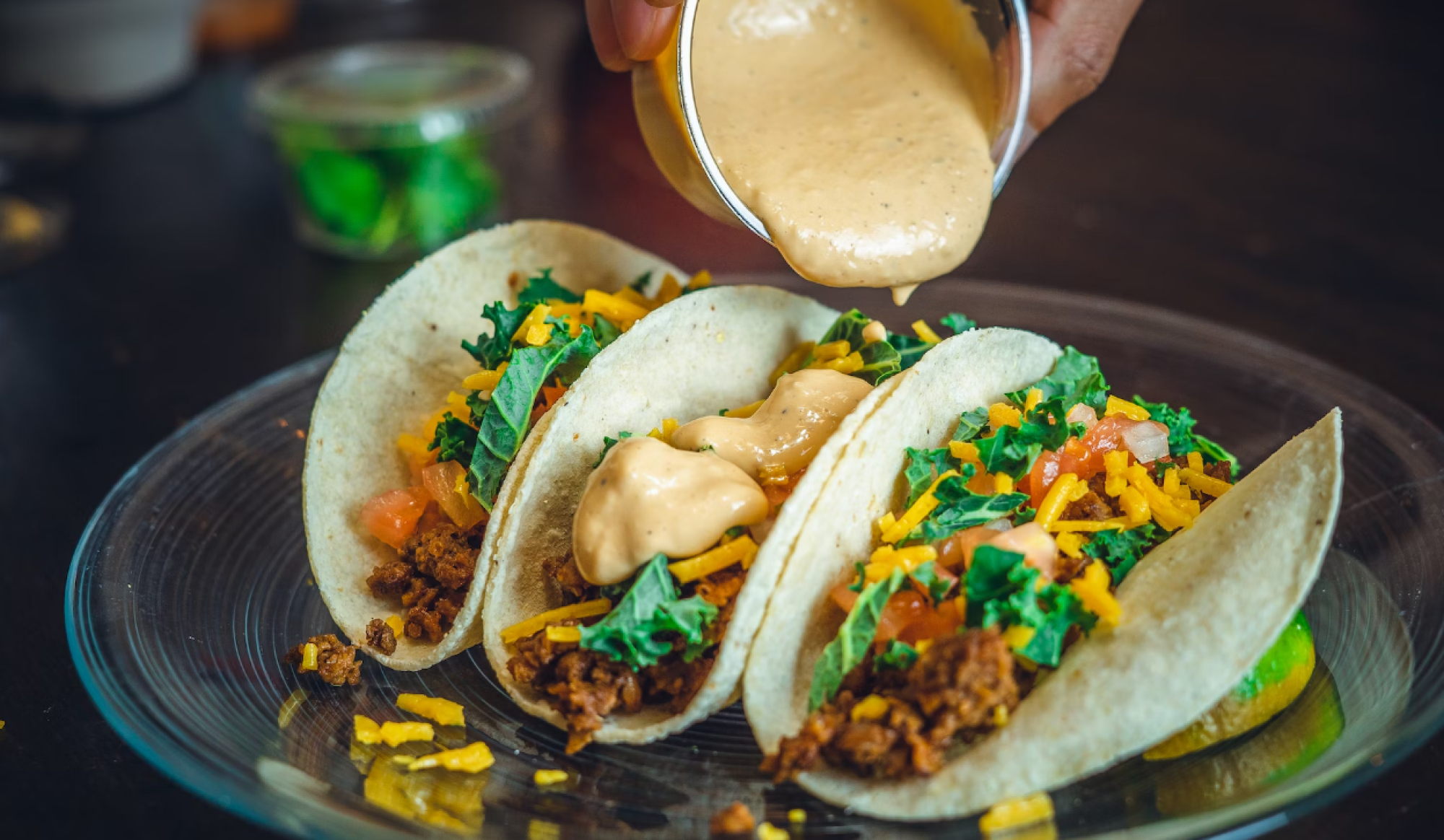 Three tacos filled with meat, cheese, lettuce, and tomato are being topped with a creamy sauce on a glass plate.