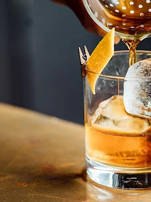 A cocktail is being poured into a glass with ice and garnished with a citrus peel, set on a tabletop.