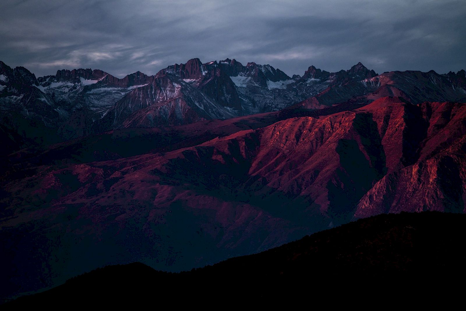A rugged mountain range with snow-capped peaks, illuminated by a dramatic red sunset under a cloudy sky.