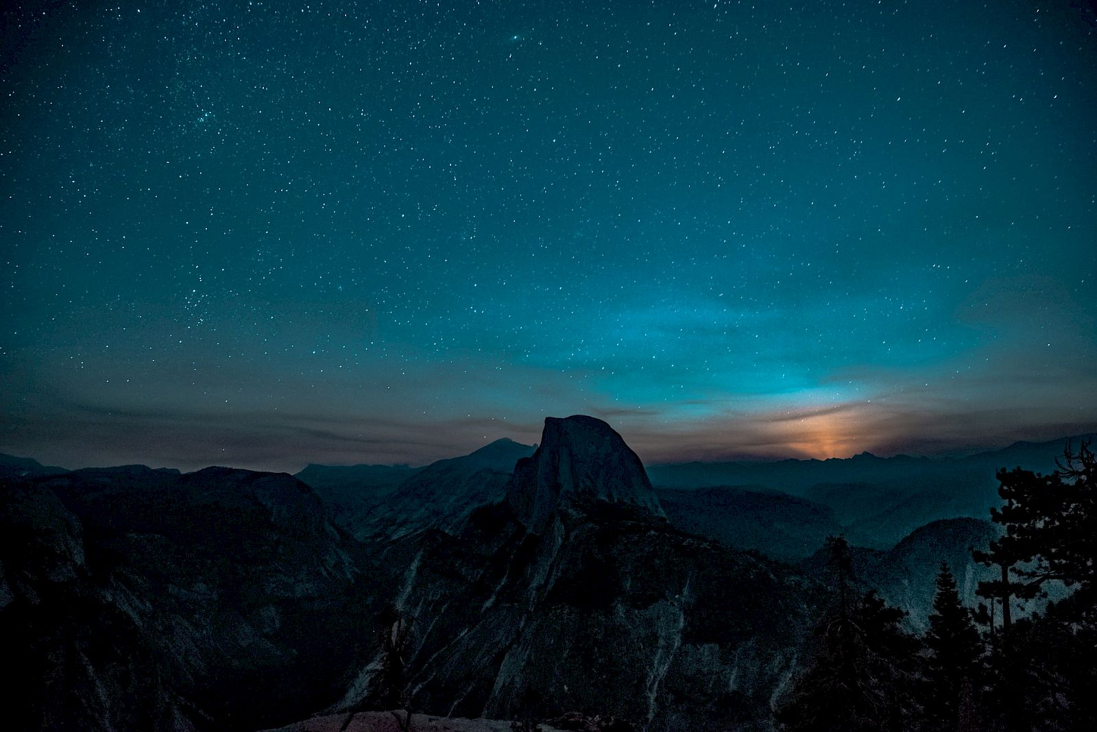 A mountainous landscape under a starry night sky with a glowing horizon, silhouetted trees, and a prominent peak in the foreground.