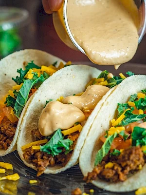 Three tacos with ground meat, lettuce, cheese, and sauce being poured on top on a plate.