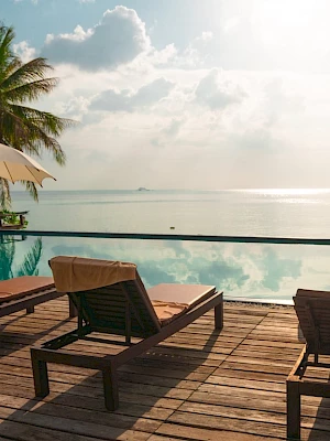 Poolside view with lounge chairs, umbrellas, and a tranquil sea under a cloudy sky. Palm trees frame the relaxing scene perfectly.