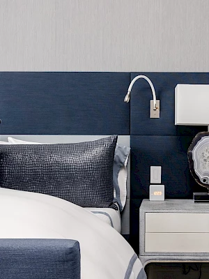 A modern bedroom with a navy headboard, bed, pillows, a white lamp on a nightstand, and a sleek wall-mounted reading light.