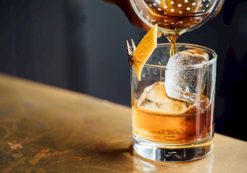A cocktail is being poured into a glass with large ice cubes, garnished with a twist of citrus peel.