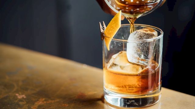 A cocktail is being poured into a glass with large ice cubes, garnished with a twist of citrus peel.