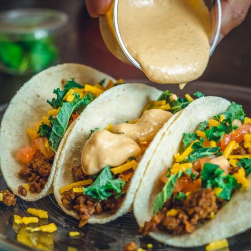 Three tacos filled with meat, cheese, tomatoes, and greens being topped with a creamy sauce, served on a plate.