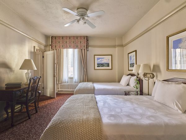 The image shows a hotel room with two beds, a desk, lamps, and framed art on the walls, featuring beige and neutral tones.