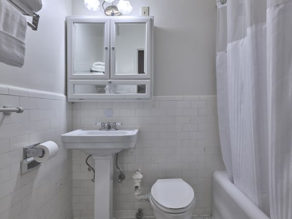A small, clean bathroom with a pedestal sink, toilet, mirrored cabinet, and a bathtub with a shower curtain. White tiles cover the floor and walls.
