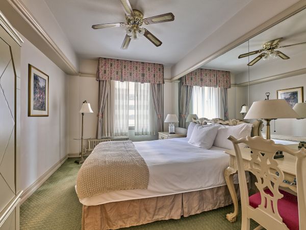 A well-decorated bedroom with a double bed, mirrored wall, ceiling fan, and lamps, featuring floral curtains and carpeted flooring.