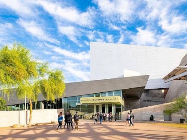 The image shows a modern building complex with people walking outside under a bright blue sky with clouds.
