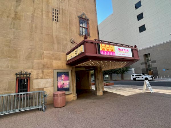 A vintage theater entrance with ornate architecture and a marquee sign. Posters are displayed, and nearby buildings are visible.