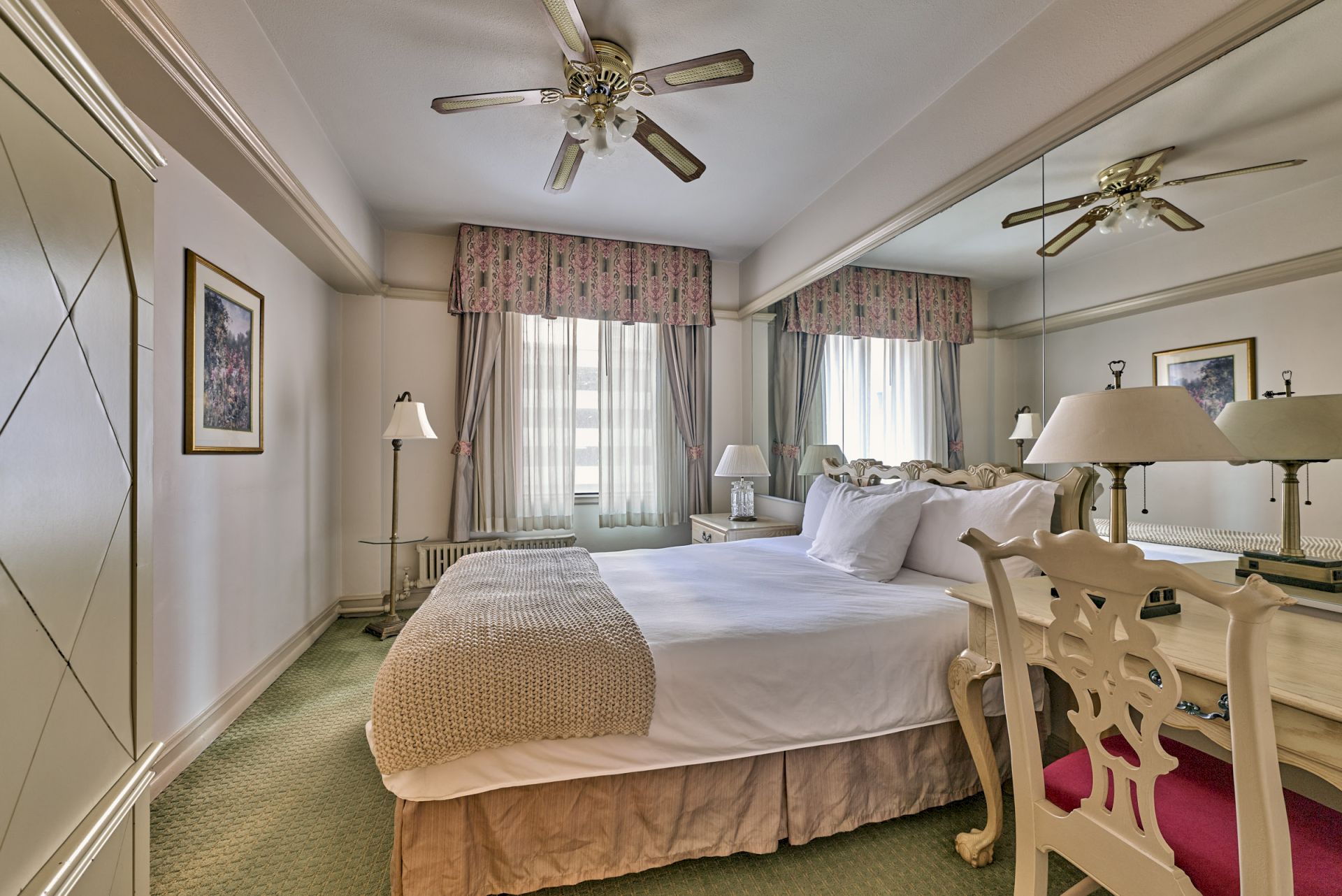 A neatly arranged bedroom with a double bed, ceiling fans, lamps, artwork on the walls, and mirrored wardrobe doors.
