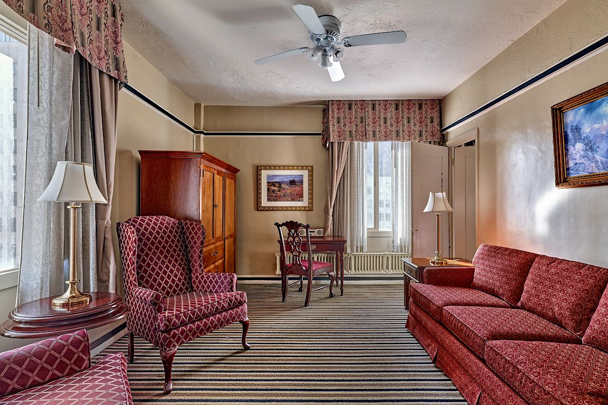 A cozy room features patterned chairs, a sofa, a wooden cabinet, a small table, and striped carpet, with paintings and curtains. Ceiling fan above.