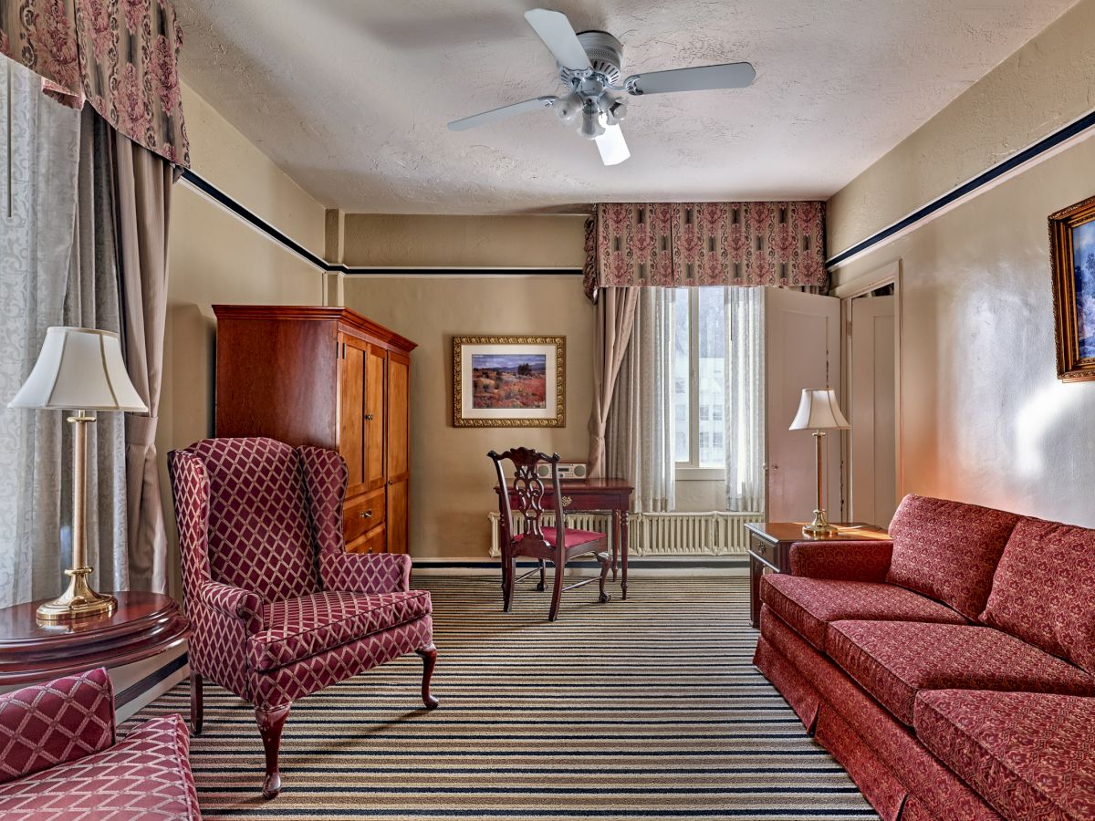 A cozy room features patterned chairs, a sofa, a wooden cabinet, a small table, and striped carpet, with paintings and curtains. Ceiling fan above.