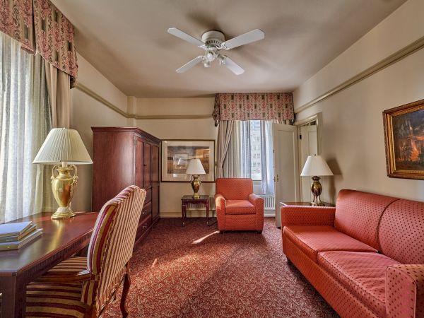 The image shows a hotel room with a desk, chairs, a couch, lamps, and a ceiling fan. There's carpet flooring and artwork on the wall.