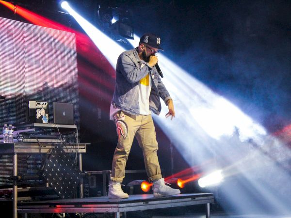 A performer is rapping on stage with dynamic lighting and DJ equipment in the background, creating an energetic atmosphere.