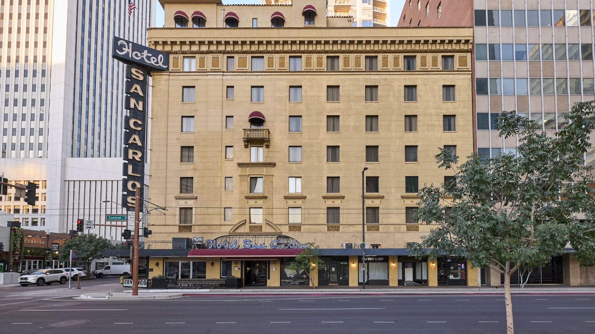 A historic multi-story hotel building is flanked by modern structures, with a "Hotel San Carlos" neon sign facing the street.