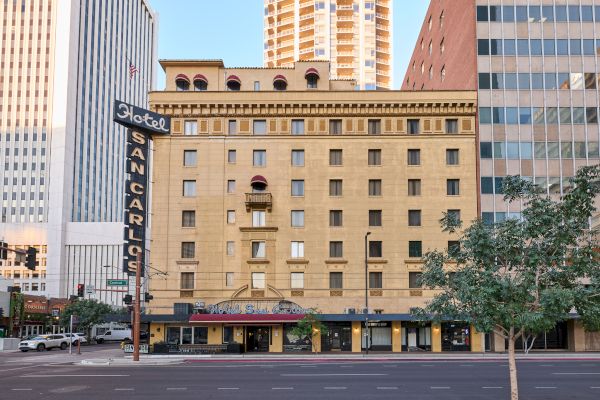 A historic hotel building with a vintage sign, located in an urban setting surrounded by skyscrapers and trees, viewed from the street.