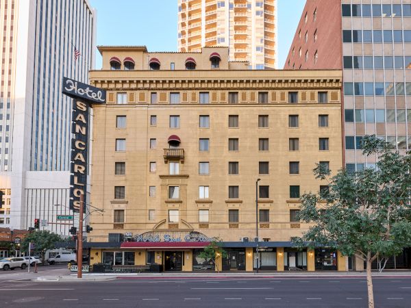 A historic hotel building with a vintage sign, located in an urban setting surrounded by skyscrapers and trees, viewed from the street.