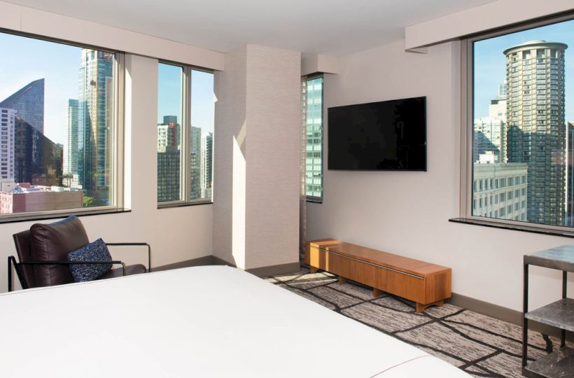 A modern hotel room with large windows showing a cityscape, a wall-mounted TV, a bed, an armchair, and a small table.