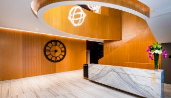 The image shows a modern lobby with a marble desk, wooden accents, a large clock, geometric lighting, and flowers in a vase.