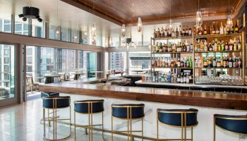 A modern bar with stylish seating, a well-stocked liquor display, and large windows offering a city skyline view.
