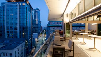 A modern balcony with seating overlooks a cityscape featuring tall buildings at dusk, adjacent to a well-lit interior space.