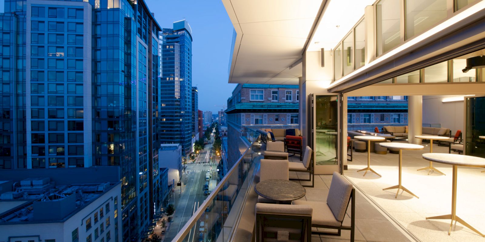 A modern balcony overlooks a city street at dusk, with tall buildings and a seating area with tables and chairs.