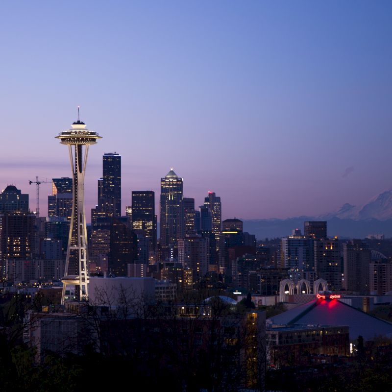A cityscape featuring the Space Needle at dusk with a mountain in the background and a purple sky.