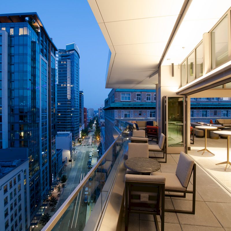 The image shows a modern rooftop patio with chairs and tables, overlooking a cityscape with tall buildings and streetlights at dusk.