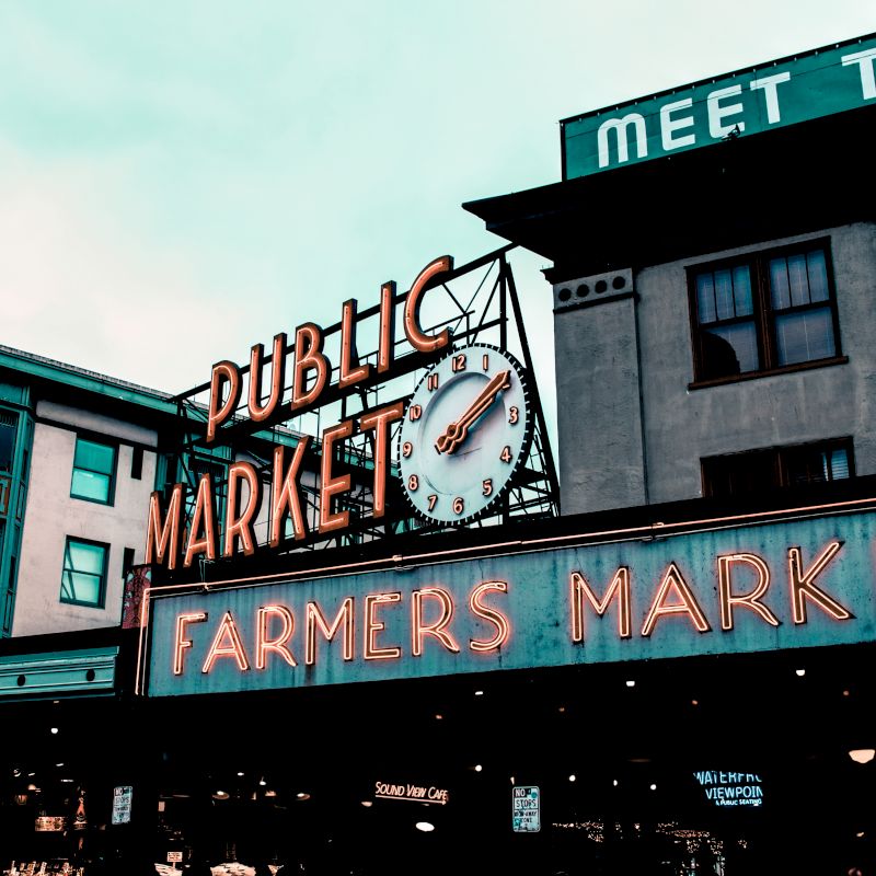 The image shows a public market sign and a clock, with "Farmers Market" in neon, likely at a bustling urban location.