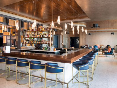 A modern bar with stylish pendant lights, a stocked liquor shelf, and blue cushioned bar stools in an open seating area.