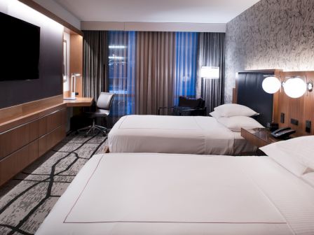 The image shows a modern hotel room with two double beds, a wall-mounted TV, a desk with a chair, and decorative lighting.