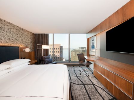 A modern hotel room with a large bed, desk, chair, and TV. The window offers a cityscape view.
