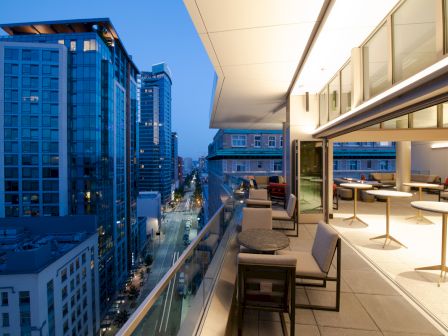 Modern cityscape with glass buildings and a stylish balcony overlooking a busy street at dusk, featuring tables and chairs.